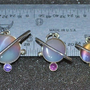 May include: Three silver charm pendants with a planet design. Each pendant features a round, iridescent glass sphere with a silver ring around it. The spheres are in shades of blue, purple, and yellow. The pendants are hanging from silver chains.