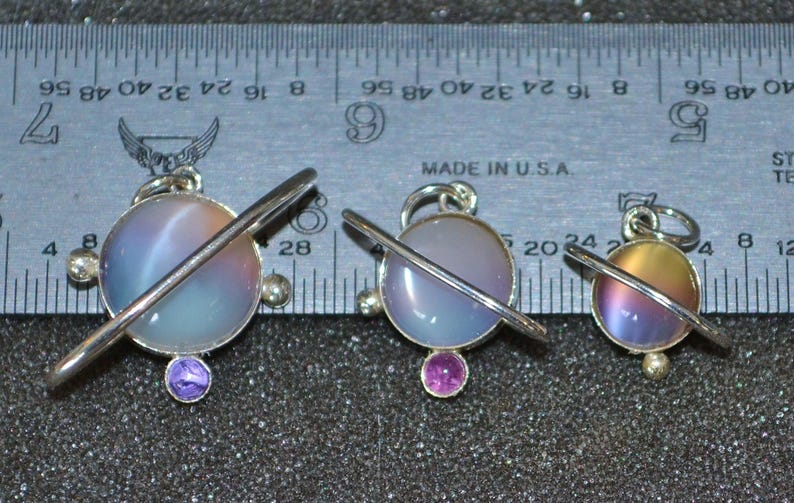 May include: Three silver charm pendants with a planet design. Each pendant features a round, iridescent glass sphere with a silver ring around it. The spheres are in shades of blue, purple, and yellow. The pendants are hanging from silver chains.