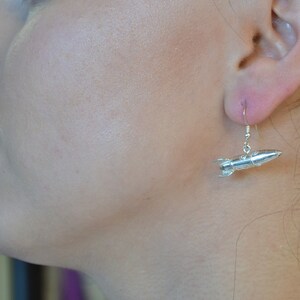 May include: Silver rocket ship dangle earrings. The rocket ship is detailed with a pointed nose and fins.
