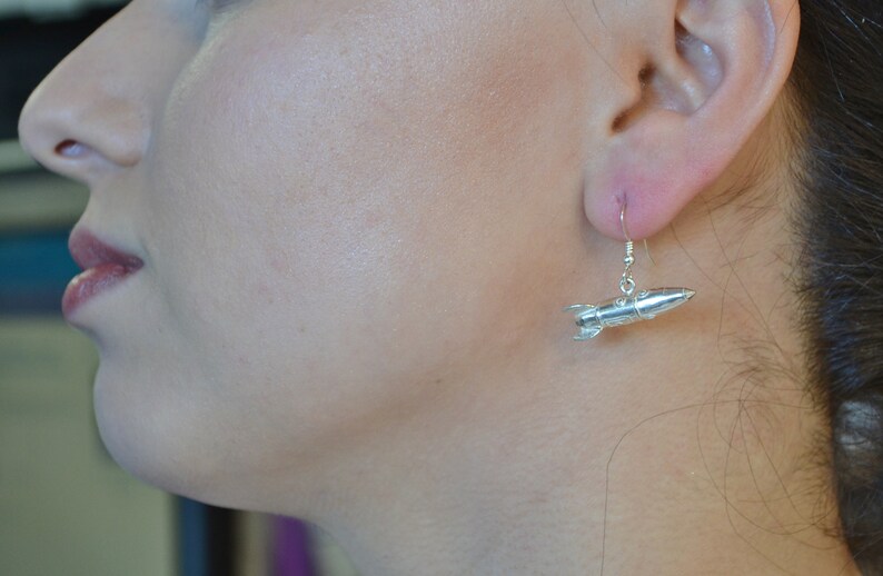 May include: Silver rocket ship dangle earrings. The rocket ship is detailed with a pointed nose and fins.