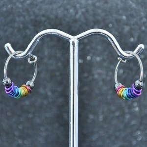 May include: A pair of silver hoop earrings with rainbow colored beads. The earrings are on a silver stand.