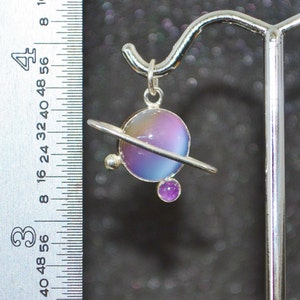 May include: A silver pendant featuring a purple and blue iridescent glass sphere with a silver ring around it, representing a planet. A smaller purple gemstone is attached to the bottom of the ring.