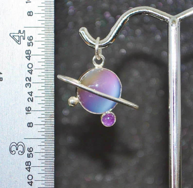 May include: A silver pendant featuring a purple and blue iridescent glass sphere with a silver ring around it, representing a planet. A smaller purple gemstone is attached to the bottom of the ring.