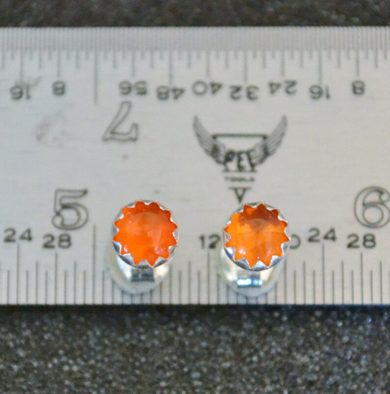 May include: A pair of orange gemstone cufflinks with a silver setting. The cufflinks are measured against a ruler.