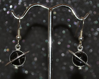 Puede incluir: Un par de pendientes de plata con piedras negras en forma de estrella y un anillo de plata alrededor de cada piedra, que se asemeja a un planeta con un sistema de anillos. Los pendientes están colgados de un soporte de plata.