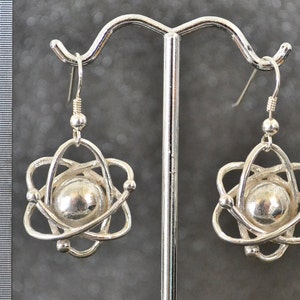Pu&ograve; includere: Orecchini in argento a forma di atomo con un centro sferico ed elettroni in orbita. Gli orecchini sono appesi a un gancio in argento.