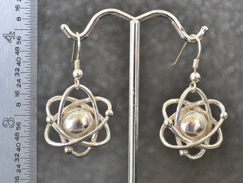 Pu&ograve; includere: Orecchini in argento a forma di atomo con un centro sferico ed elettroni in orbita. Gli orecchini sono appesi a un gancio in argento.