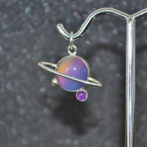 May include: A silver charm necklace featuring a purple and pink iridescent glass sphere with a silver ring around it, representing a planet. A smaller purple gemstone is attached to the bottom of the ring.