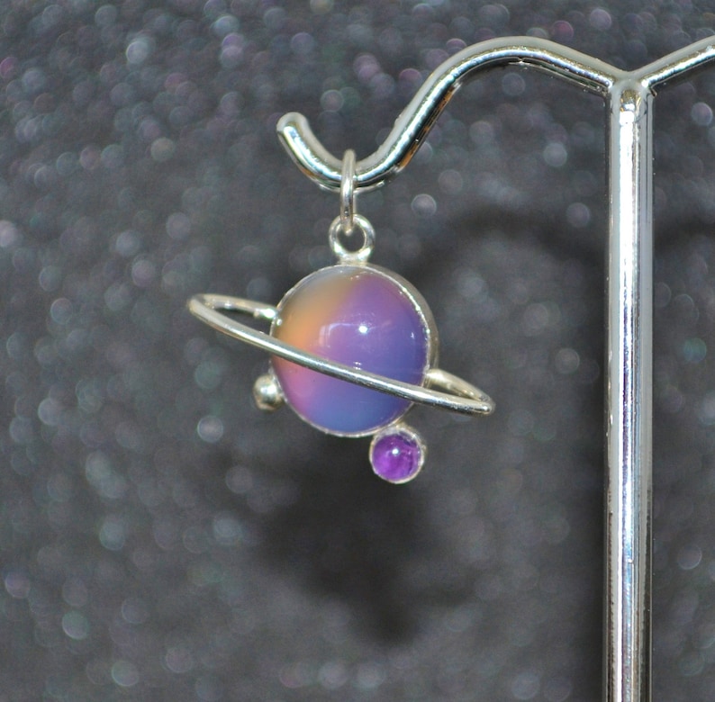 May include: A silver charm necklace featuring a purple and pink iridescent glass sphere with a silver ring around it, representing a planet. A smaller purple gemstone is attached to the bottom of the ring.