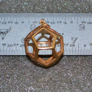 Pode incluir: Um pingente geom&eacute;trico de cor cobre com um design aberto e complexo. O pingente &eacute; suspenso por um pequeno la&ccedil;o. Uma r&eacute;gua com medidas em polegadas est&aacute; no fundo. O pingente tem aproximadamente 3,8 cm de di&acirc;metro.
