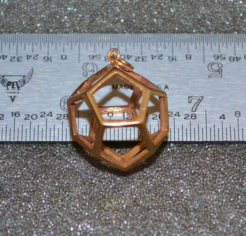Pode incluir: Um pingente geom&eacute;trico de cor cobre com um design aberto e complexo. O pingente &eacute; suspenso por um pequeno la&ccedil;o. Uma r&eacute;gua com medidas em polegadas est&aacute; no fundo. O pingente tem aproximadamente 3,8 cm de di&acirc;metro.