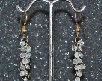 May include: A pair of gold dangle earrings with clear crystal beads. The earrings are hanging from a silver stand.
