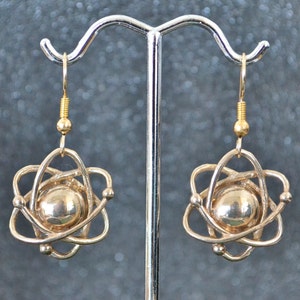 Może przedstawiać: Złote kolczyki w kształcie atomu z okrągłym środkiem i wzorem z drutu. Kolczyki wiszą na srebrnym stojaku.