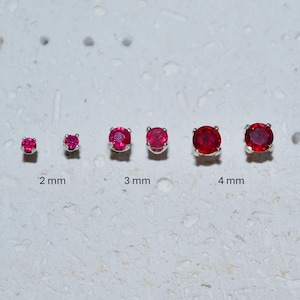 Może przedstawiać: Sześć srebrnych kolczyków sztyftów, każdy z okrągłym, jaskrawoczerwonym kamieniem szlachetnym. Kolczyki są prezentowane w różnych rozmiarach, o średnicy 2 mm, 3 mm i 4 mm. Kamienie szlachetne mają wypolerowaną, odblaskową powierzchnię.