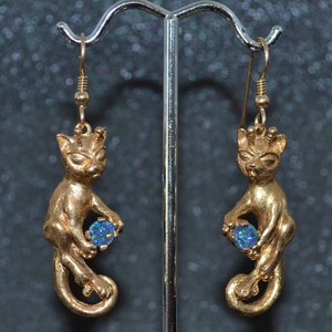 May include: Gold cat earrings with blue opal gemstones. The cats are holding the gemstones in their paws. The earrings are hanging from a silver stand.