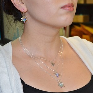 May include: A silver necklace with four layered chains and three planet pendants. Each pendant features a different colored glass orb with a silver ring around it. The necklace is worn by a person with a white shirt and black tank top.