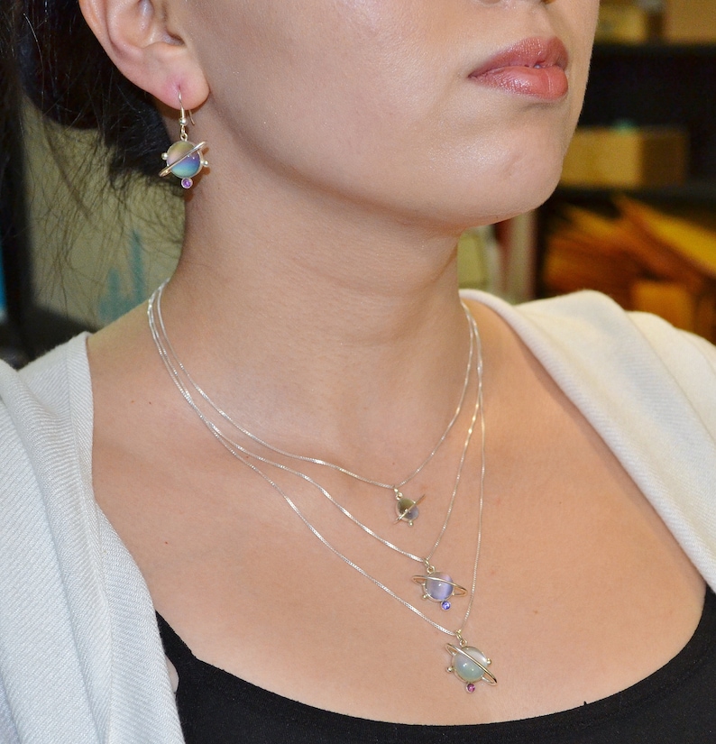 May include: A silver necklace with four layered chains and three planet pendants. Each pendant features a different colored glass orb with a silver ring around it. The necklace is worn by a person with a white shirt and black tank top.
