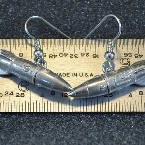 May include: A pair of silver rocket ship earrings with a hook closure. The earrings are about 1.5 inches long and are shown next to a ruler for size reference.