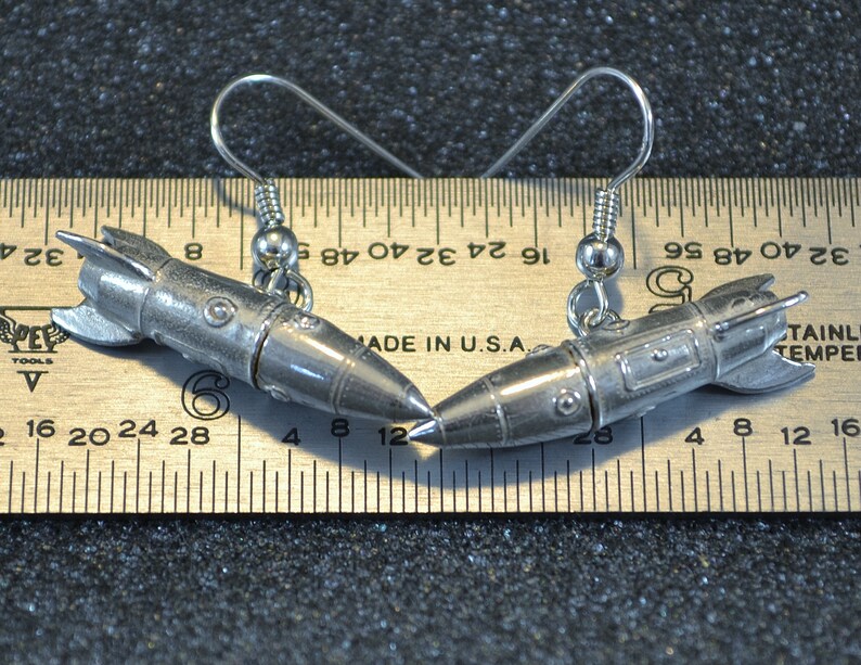 May include: A pair of silver rocket ship earrings with a hook closure. The earrings are about 1.5 inches long and are shown next to a ruler for size reference.