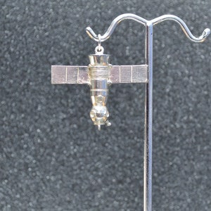May include: A silver charm in the shape of a space station with solar panels. The charm is hanging from a silver stand.