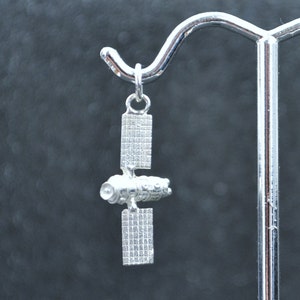 Puede incluir: Encanto de plata de una estación espacial con un diseño detallado. El encanto cuelga de una cadena de plata.