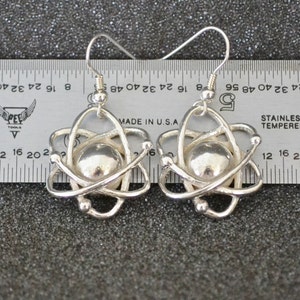 Pu&ograve; includere: Un paio di orecchini a forma di atomo in argento con una piccola sfera d'argento al centro di ogni atomo. Gli orecchini sono appesi a un righello con il testo "MADE IN U.S.A" e "STAINLESS TEMPERED" visibile.