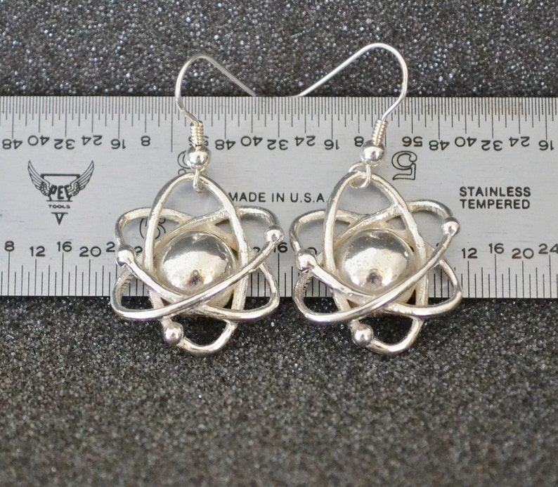 Pu&ograve; includere: Un paio di orecchini a forma di atomo in argento con una piccola sfera d'argento al centro di ogni atomo. Gli orecchini sono appesi a un righello con il testo "MADE IN U.S.A" e "STAINLESS TEMPERED" visibile.