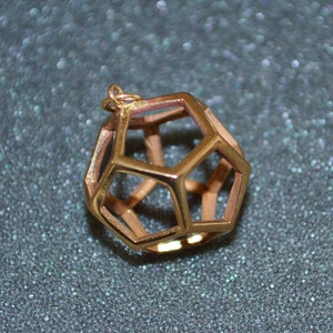 Pode incluir: Um pendente geom&eacute;trico dourado com um design oco e aberto. O pendente tem a forma de um poliedro, com m&uacute;ltiplas faces pentagonais e hexagonais. O pendente tem uma pequena argola no topo para uma corrente.