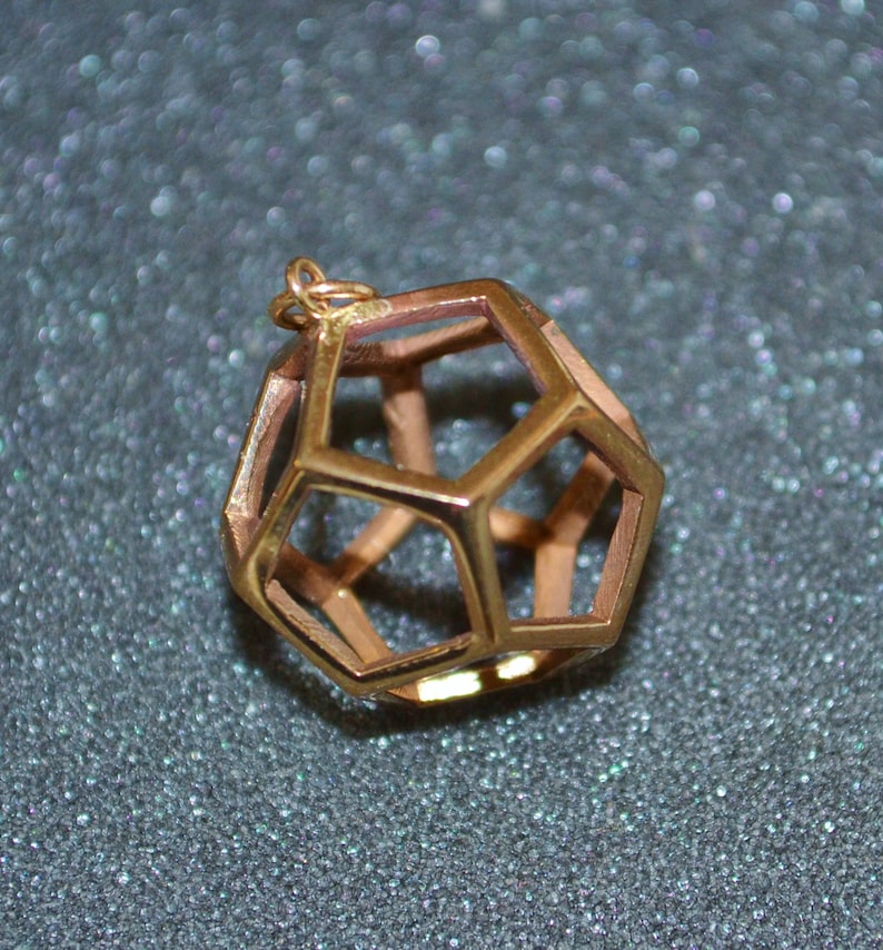 Pode incluir: Um pendente geom&eacute;trico dourado com um design oco e aberto. O pendente tem a forma de um poliedro, com m&uacute;ltiplas faces pentagonais e hexagonais. O pendente tem uma pequena argola no topo para uma corrente.