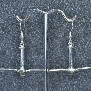 May include: Silver-toned lighthouse earrings. Each earring features a detailed lighthouse design with a vertical column and horizontal extensions. The earrings hang from a simple hook. The background is a textured, dark gray surface.