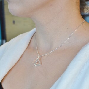 Puede incluir: Un collar de plata con un colgante geom&eacute;trico en forma de pir&aacute;mide. El colgante est&aacute; hecho de alambre fino y tiene un centro hueco.