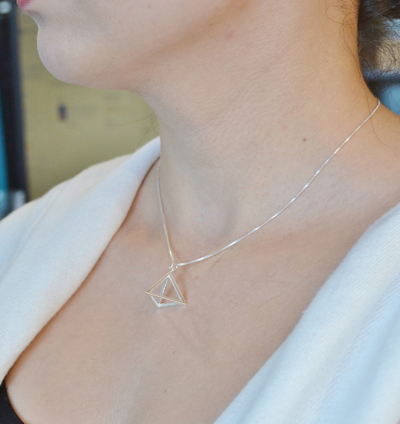 Puede incluir: Un collar de plata con un colgante geom&eacute;trico en forma de pir&aacute;mide. El colgante est&aacute; hecho de alambre fino y tiene un centro hueco.