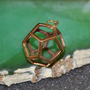 Pode incluir: Um pendente geom&eacute;trico dourado, com uma estrutura oca e multifacetada. Possui uma pequena argola para uma corrente. O fundo &eacute; uma mistura de verde e castanho. O design do pendente &eacute; intrincado.