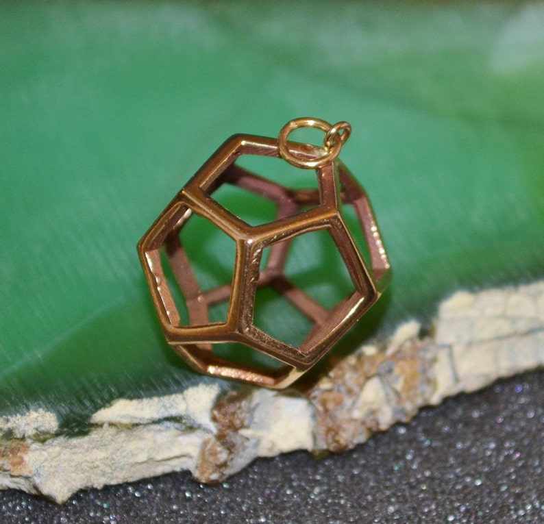 Pode incluir: Um pendente geom&eacute;trico dourado, com uma estrutura oca e multifacetada. Possui uma pequena argola para uma corrente. O fundo &eacute; uma mistura de verde e castanho. O design do pendente &eacute; intrincado.