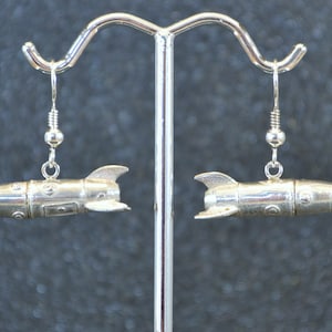 May include: A pair of silver rocket ship earrings hanging from a display stand. The earrings are detailed with small rivets and have a pointed nose.