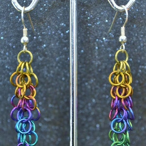 May include: A pair of rainbow colored chain earrings hanging from silver hooks. The earrings are made of small metal rings that are linked together to form a chain. The rings are graduated in size, with the smallest rings at the bottom and the largest rings at the top. The earrings are a vibrant mix of colors, including yellow, orange, red, purple, blue, and green.