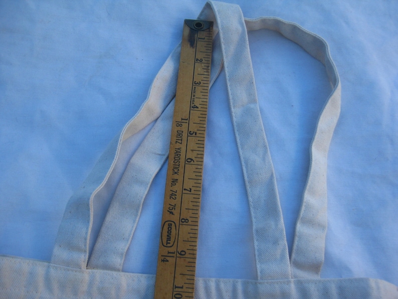 May include: A cream-colored canvas tote bag with matching straps. A wooden ruler, marked in inches, is placed vertically against the bag, showing measurements. The ruler is marked with the text "18 DRITZ YARDSTICK No. 742 75f SCOVILL".