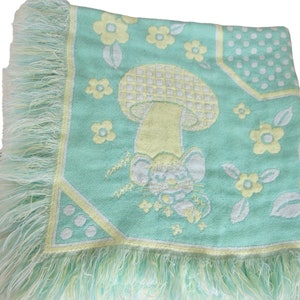 Puede incluir: Una manta tejida verde y amarilla con un borde de flecos. La manta presenta un ratón de dibujos animados y un hongo con un patrón a cuadros.