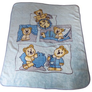 May include: A light blue baby blanket featuring cartoon teddy bears in various sleeping poses. The blanket has a soft texture and a border. The bears are depicted with pillows, pajamas, and a clock, creating a bedtime theme.