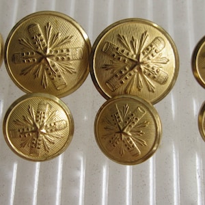 May include: A collection of nine gold-colored buttons. The buttons are round with a detailed embossed design featuring a starburst pattern with crossed elements. The buttons vary in size, with some larger than others.