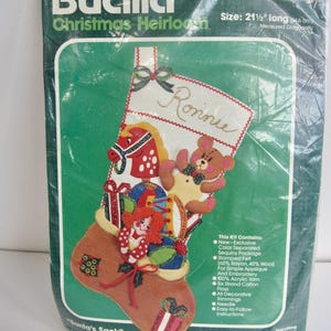 Puede incluir: Un kit de medias de punto de aguja con joyas de Bucilla Christmas Heirloom, con una media de fieltro con un caballo de balancín rojo y blanco, un oso de peluche y una muñeca. La media mide 55 cm de largo e incluye lentejuelas, hilo, hilo de bordar e instrucciones. El kit está empaquetado en una bolsa de plástico transparente con fondo verde.