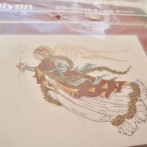 May include: A cross stitch pattern featuring a white angel with a red and green robe, holding a star. The pattern is titled "Christmas Cross Stitch".