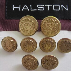 May include: Eight circular gold-toned metal buttons featuring an embossed letter H surrounded by a laurel wreath design. A black fabric label with the white embroidered text HALSTON is positioned above the buttons on a dark maroon fabric background.