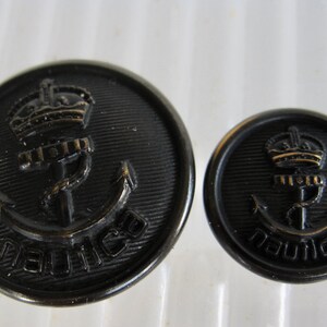 May include: Two black nautical buttons with an anchor, crown, and the word "nautica". The buttons are round with a ridged edge. One button is larger than the other.