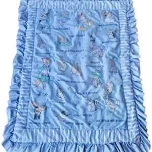 Puede incluir: Una manta para bebé azul y blanca con un diseño acolchado con personajes de los libros de Beatrix Potter. Los personajes incluyen Peter Rabbit, Mrs. Rabbit, Jemima Puddle-Duck, Mr. Tod y Squirrel Nutkin. La manta tiene un borde con volantes.