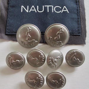 Puede incluir: Un conjunto de ocho botones redondos de metal plateado con un diseño de ancla en relieve y el texto NAUTICA. Los botones están dispuestos frente a una etiqueta de tela azul marino oscuro con el texto NAUTICA bordado en blanco.
