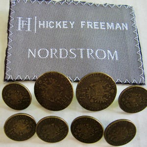 May include: A rectangular grey fabric label with white embroidered text reading Hickey Freeman and Nordstrom, featuring a zigzag stitched border. Below the label are eight round, antique brass-toned metal buttons with an embossed eagle emblem.