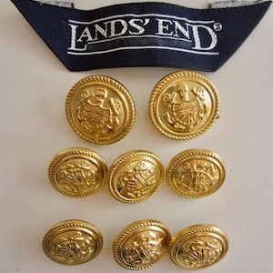 Puede incluir: Ocho botones de color dorado con un diseño en relieve de un escudo y anclas. Los botones tienen un borde de cuerda. Los botones están dispuestos en dos filas de cuatro. Los botones están sobre un fondo blanco. El texto "LANDS' END" es visible en una etiqueta azul.