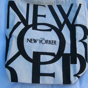 May include: Off-white tote bag with large black text spelling "NEW YORKER". The words "The New Yorker" are printed in a smaller black font below the large text. The bag is made of a light-colored fabric.