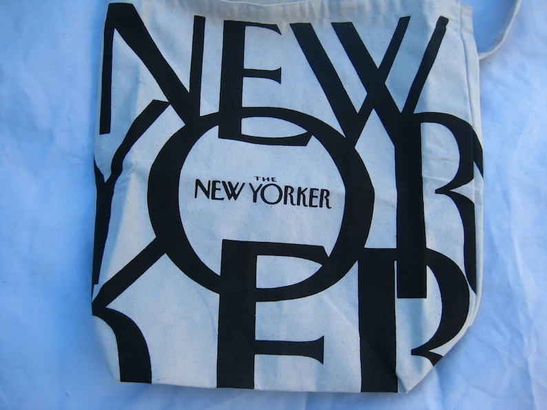 May include: Off-white tote bag with large black text spelling "NEW YORKER". The words "The New Yorker" are printed in a smaller black font below the large text. The bag is made of a light-colored fabric.